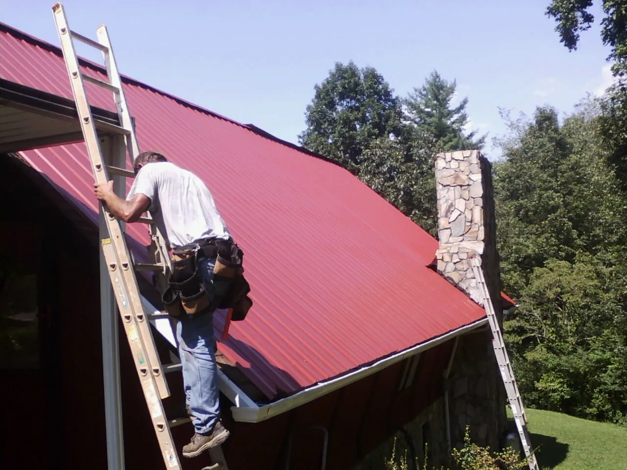Roofer on a ladder beside a red metal roof and stone chimney — Soffit & Fascia Repair by Roofing Brothers in Garfield